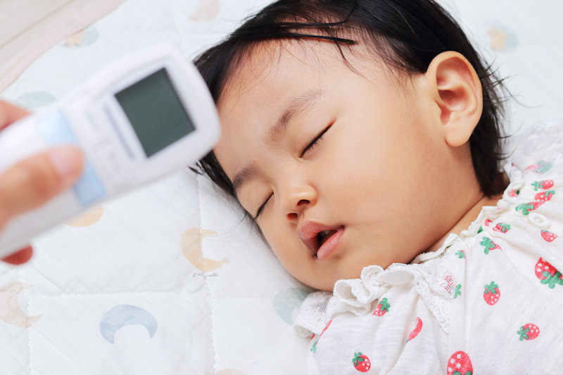 Child sleeping while a caregiver checks their temperature with a digital thermometer.